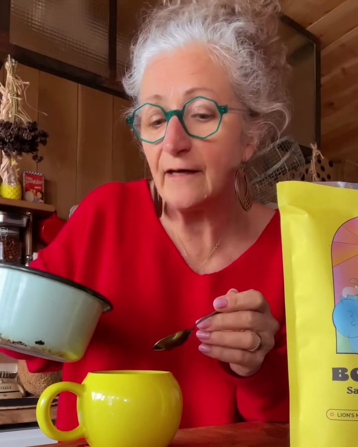 A woman pouring warm oat milk from a small saucepan into a yellow mug of Bonjour and stirring it, the pouch standing beside her on the counter